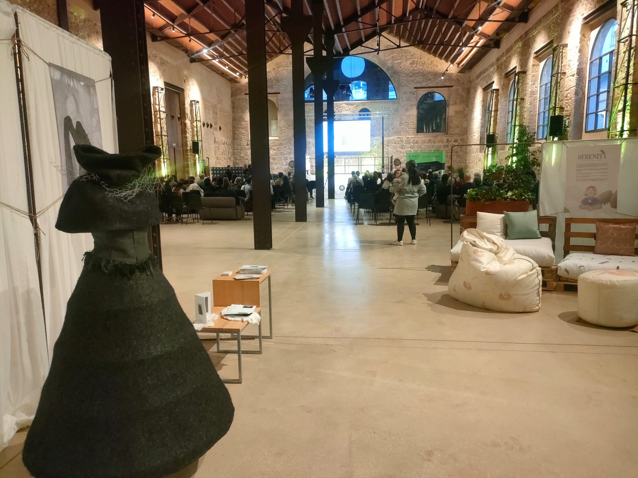 Un vestit de dol d'una proposta i els pufs d'altra, amb les presentacions de projectes al fons. 